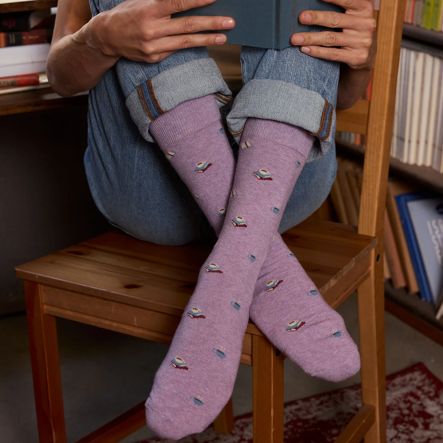 Person sitting on a wooden chair wearing purple socks with a mug and book pattern, holding a book.